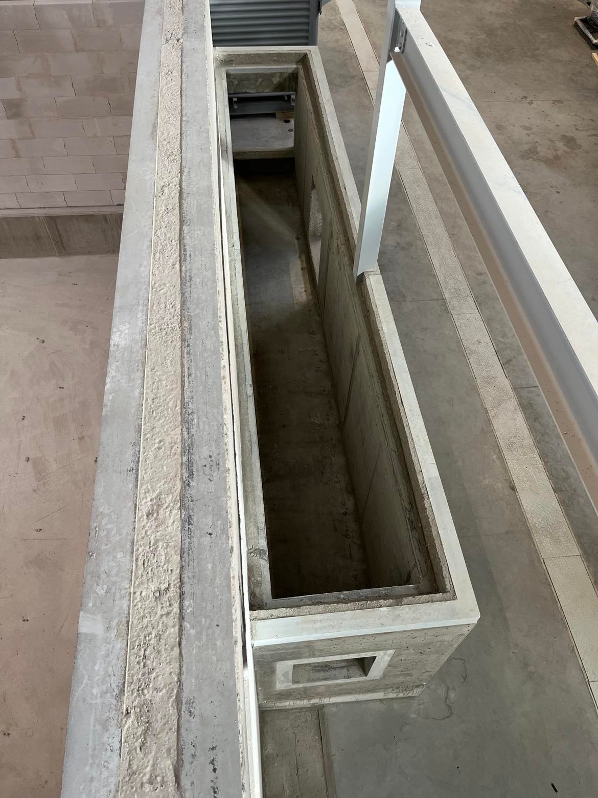 Surowy betonowy szyb z otworem rewizyjnym u dołu, widok z góry, fragment ściany z białych bloczków w tle.