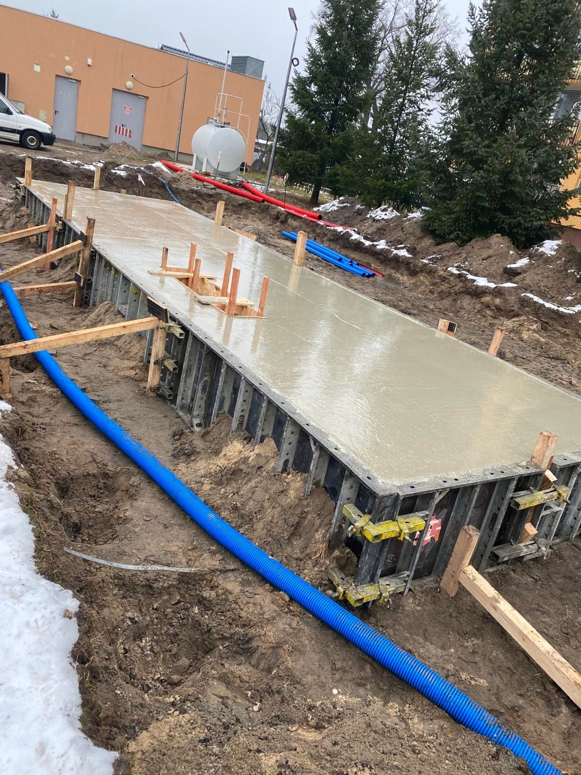 Wylany fundament z szalunkiem metalowym, widoczne podpory drewniane i niebieska rura drenażowa w wykopie, w tle budynek i drzewa iglaste.