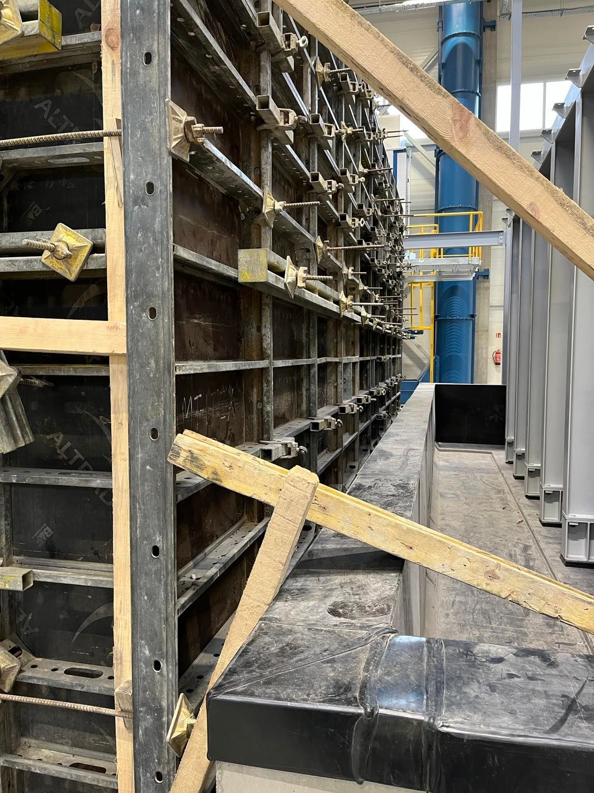 Szczegółowe ujęcie szalunku z metalowymi wzmocnieniami i drewnianymi rozpórkami, widoczne elementy konstrukcyjne w tle hali.