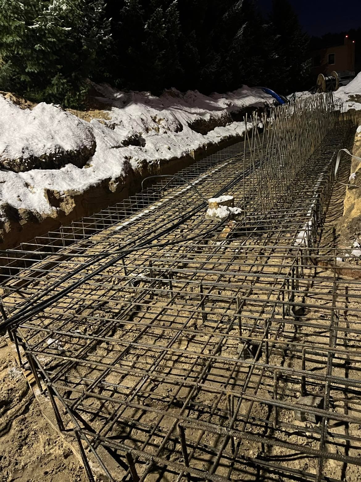 Zbrojenie pod fundament w wykopie, widoczne pręty stalowe i siatka zbrojeniowa, ziemia pokryta śniegiem, nocne oświetlenie