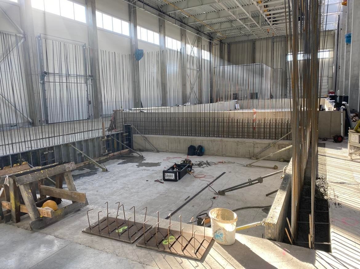 Wnętrze hali w trakcie budowy: widoczne zbrojenia ścian, szalunki, narzędzia budowlane, wiązki prętów zbrojeniowych oraz elementy drewnianej konstrukcji pomocniczej.