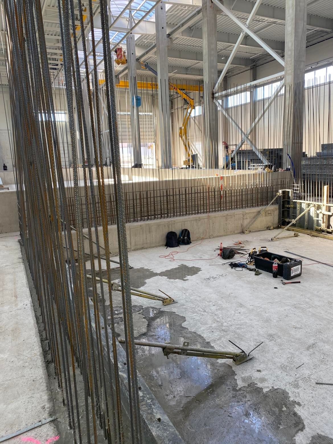 Wnętrze hali w trakcie budowy, widoczne zbrojenie ścian i słupów, na posadzce rozłożone narzędzia i sprzęt budowlany, w tle dźwig i podnośnik koszowy.