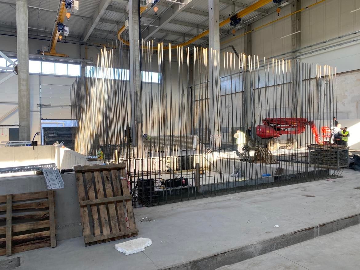 Wnętrze hali produkcyjnej z przygotowanym zbrojeniem do wylewki fundamentów, widoczny dźwig samojezdny i pracownicy budowlani w tle.