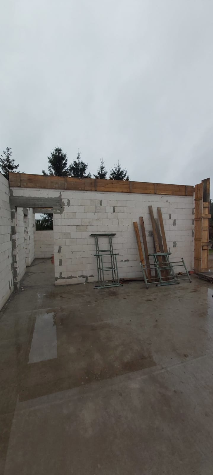 Wnętrze surowego budynku w trakcie budowy, ściany z białego bloczka, drewniane nadproża, metalowe podpory i deski leżące na podmokłej posadzce.