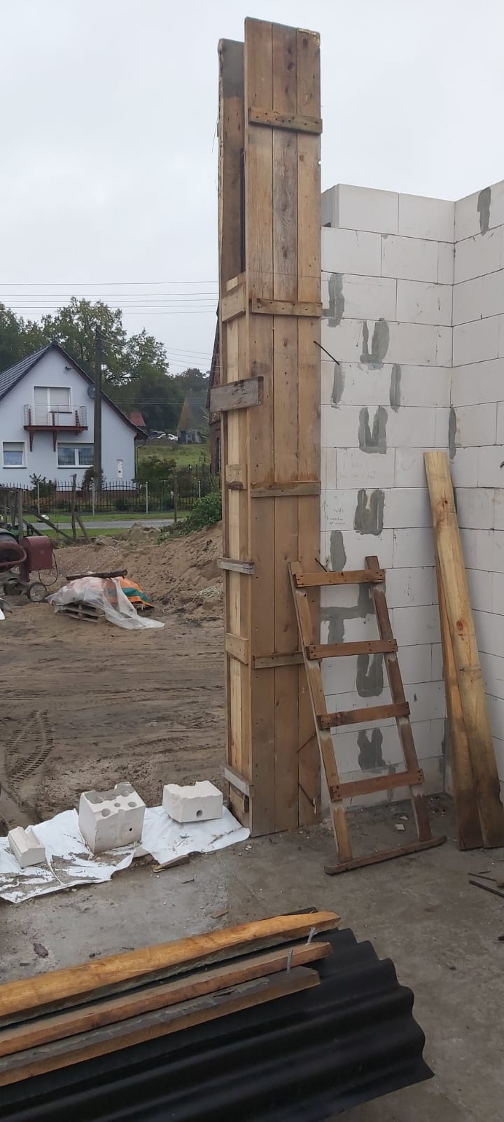Szczegół budowy domu z bloczków: drewniany szalunek kolumny narożnej, wsparty o ścianę i drewnianą drabinę, w tle dom jednorodzinny i mieszalnik do betonu.