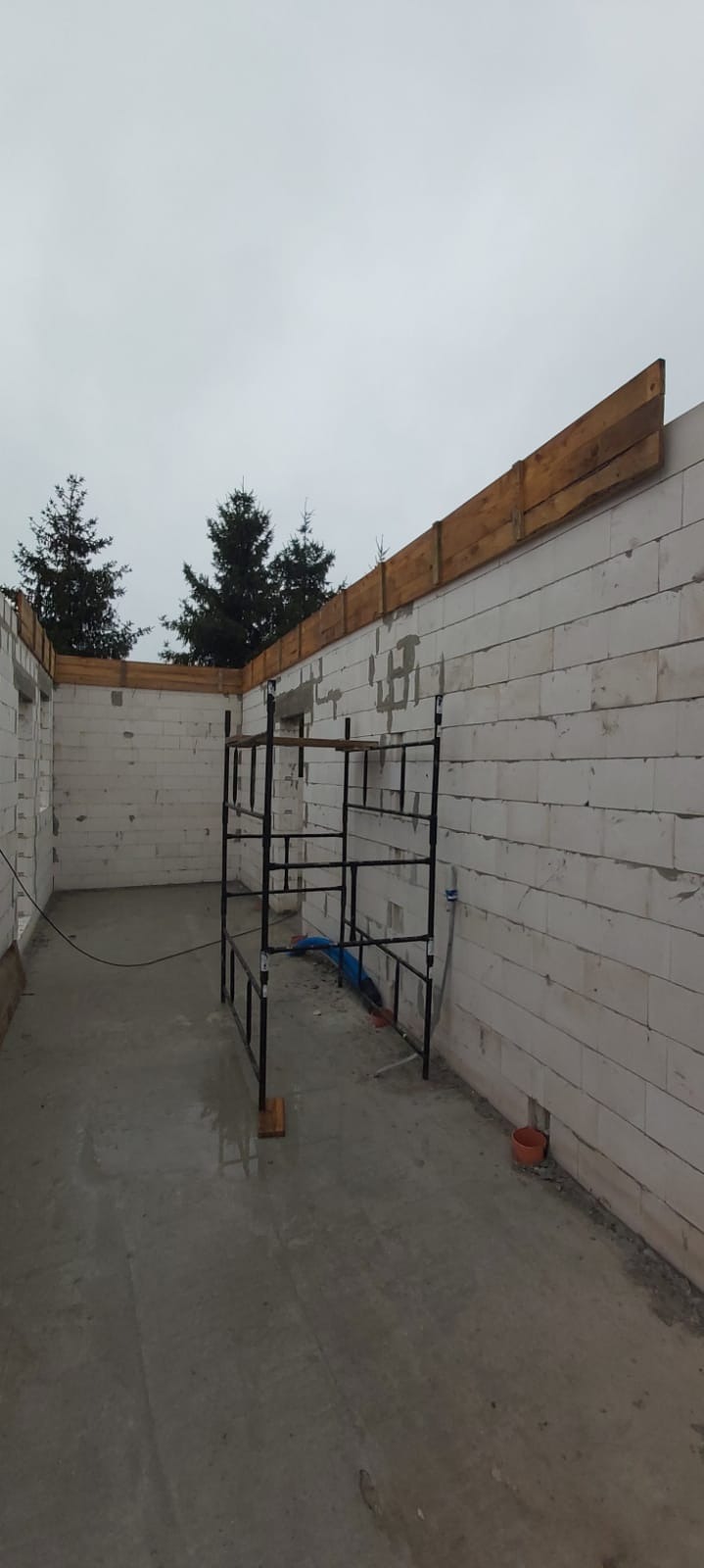 Wnętrze w trakcie budowy z białych bloczków, widoczne rusztowanie i drewniane szalunki stropowe, szare, betonowe podłoże, na zewnątrz iglaste drzewa.