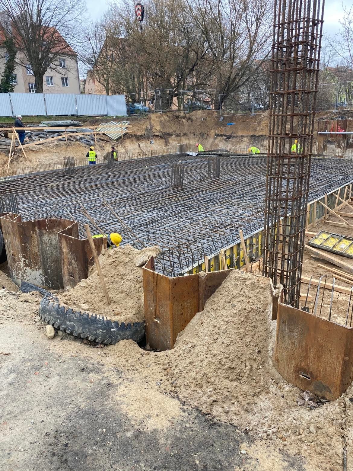 Przygotowanie zbrojenia fundamentów pod budynek murowany, widoczne stalowe pręty, szalunki, wykop ziemny oraz robotnicy w odblaskowych kamizelkach na placu budowy.