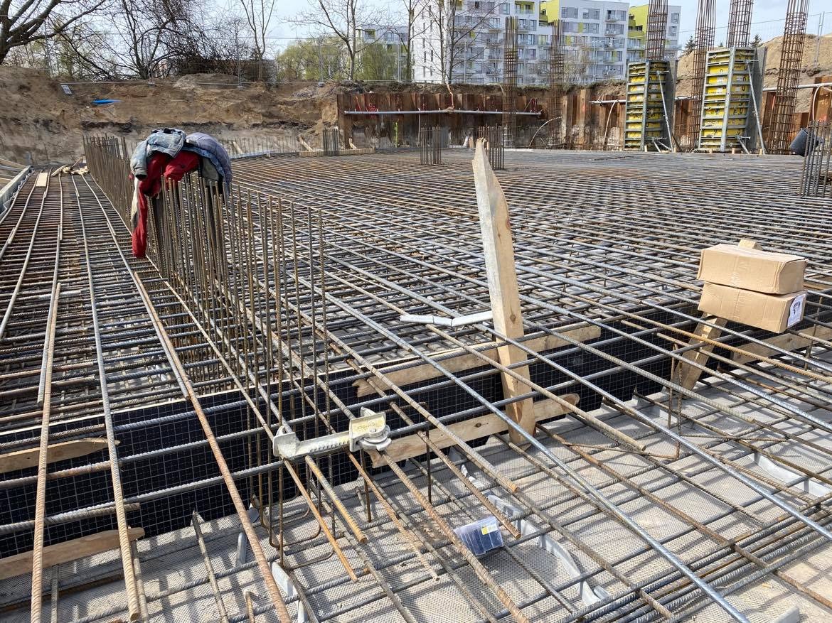 Zbrojenie płyty fundamentowej z widocznymi prętami, elementami dystansowymi i odzieżą roboczą pozostawioną na konstrukcji, w tle budynek w trakcie wznoszenia.