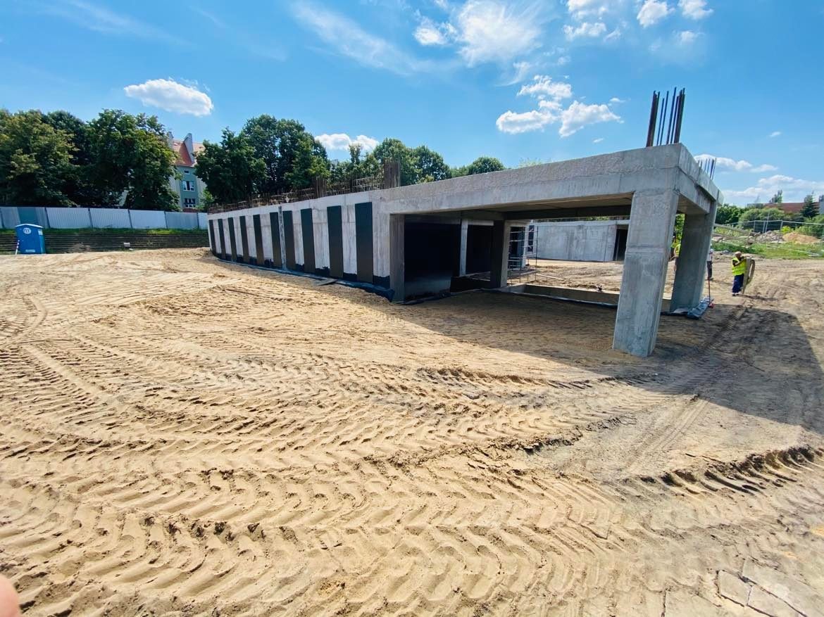 Surowa betonowa konstrukcja w trakcie budowy, widoczne zbrojenia, otoczona piaskiem z wyraźnymi śladami opon, w tle zieleń i budynek mieszkalny.