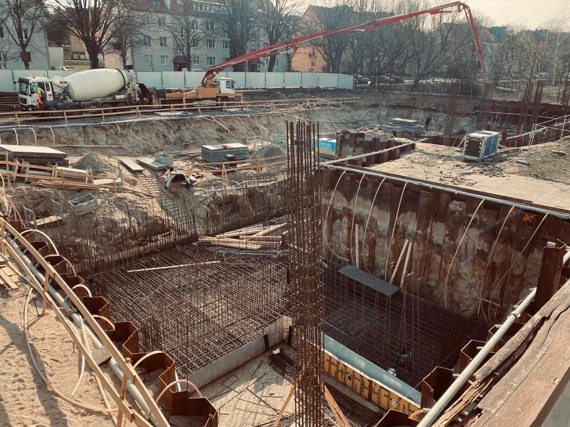 Szeroki wykop z metalowymi ścianami zabezpieczającymi, zbrojenie stalowe wewnątrz, pompa do betonu i betoniarka w tle, widoczne elementy szalunków.