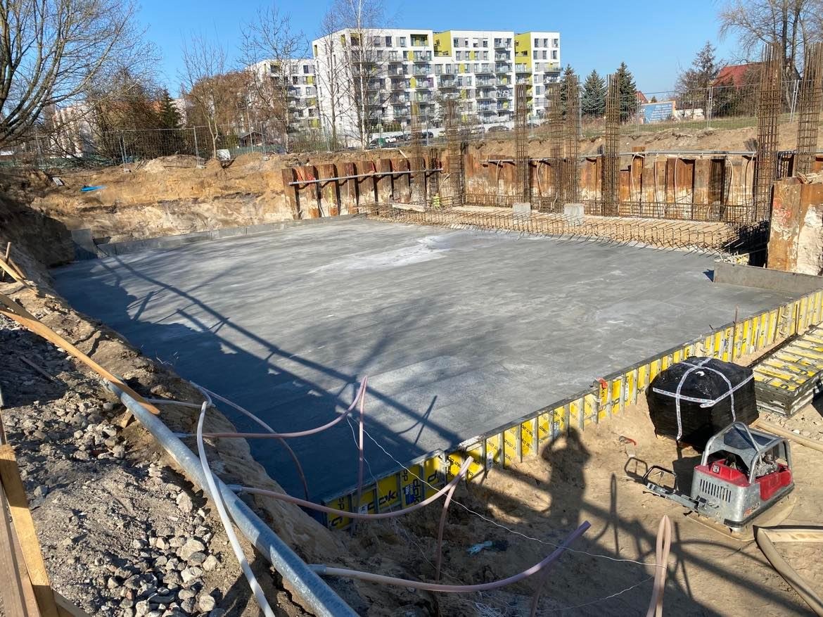 Wylana płyta fundamentowa zbrojona stalą, otoczona szalunkiem, widok z góry na plac budowy z elementami rusztowań i zabezpieczeń ziemnych, w tle budynek mieszkalny.