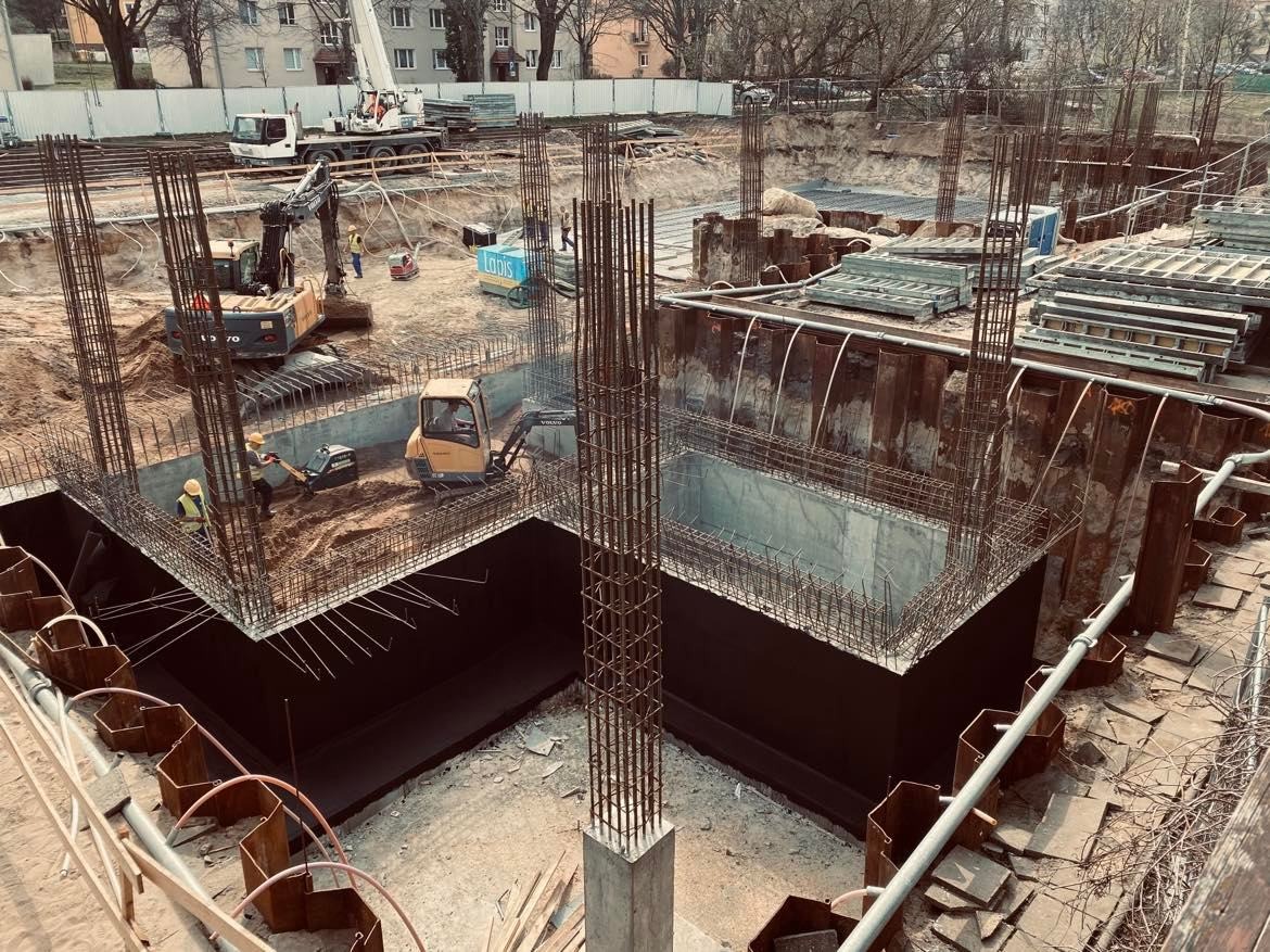 Budowa fundamentów domu: wykop z zabezpieczonymi ścianami, zbrojenie pionowe i poziome, pracujący robotnicy i maszyny budowlane w tle.