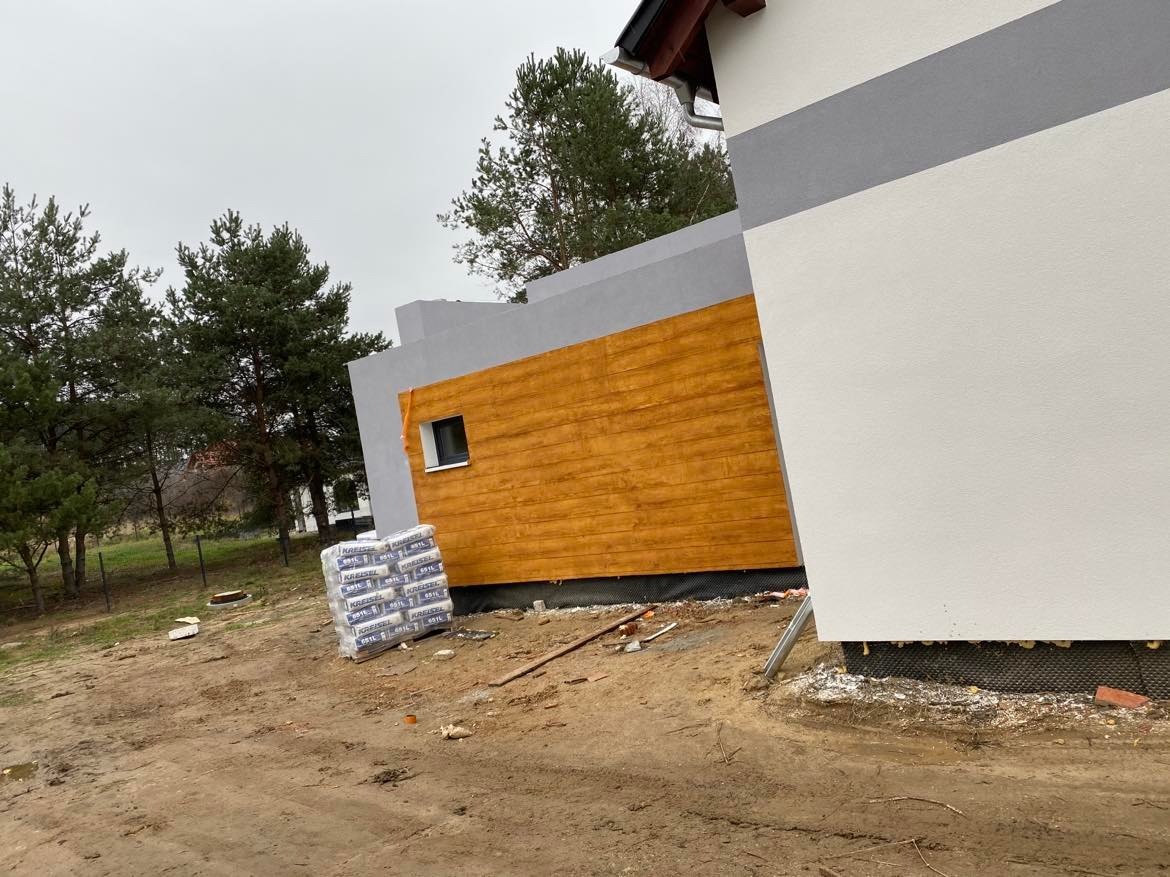 Fragment nowo wybudowanego domu z drewnianą elewacją w kolorze miodowym, widoczne worki z zaprawą budowlaną i narzędzia na placu budowy.