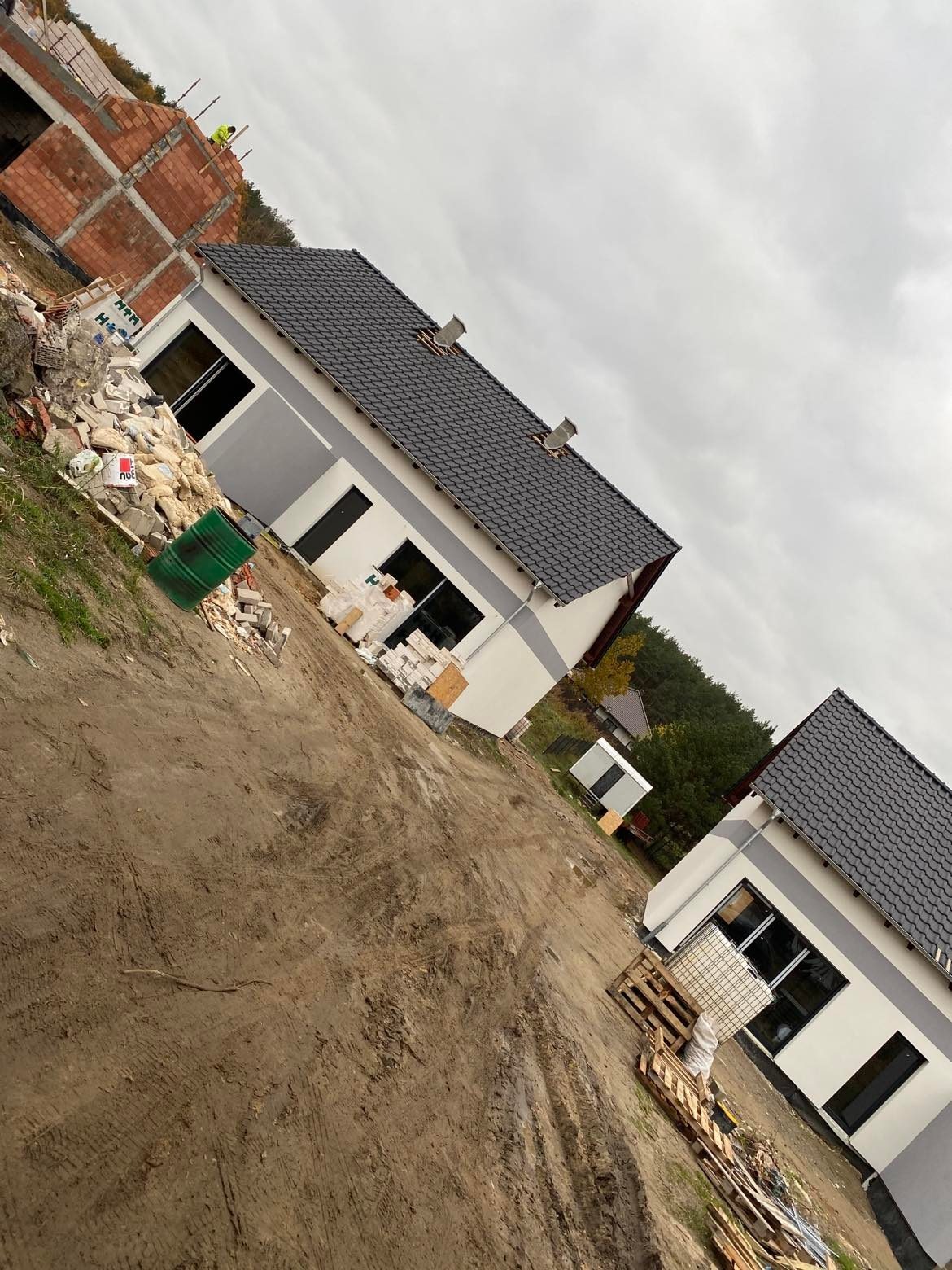 Dwa parterowe domy z szarą elewacją i ciemną dachówką w trakcie budowy, widoczne materiały budowlane, w tle konstrukcja z czerwonej cegły i zalesiony teren, pochmurne niebo.