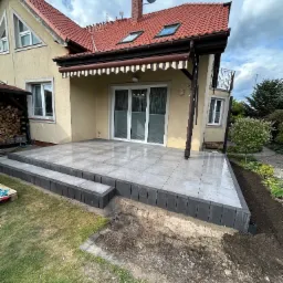 Wykończony taras z szarych płytek ceramicznych przy beżowym domu z czerwonym dachem. Widoczne schody z ciemnych kostek i fragment ogrodu.