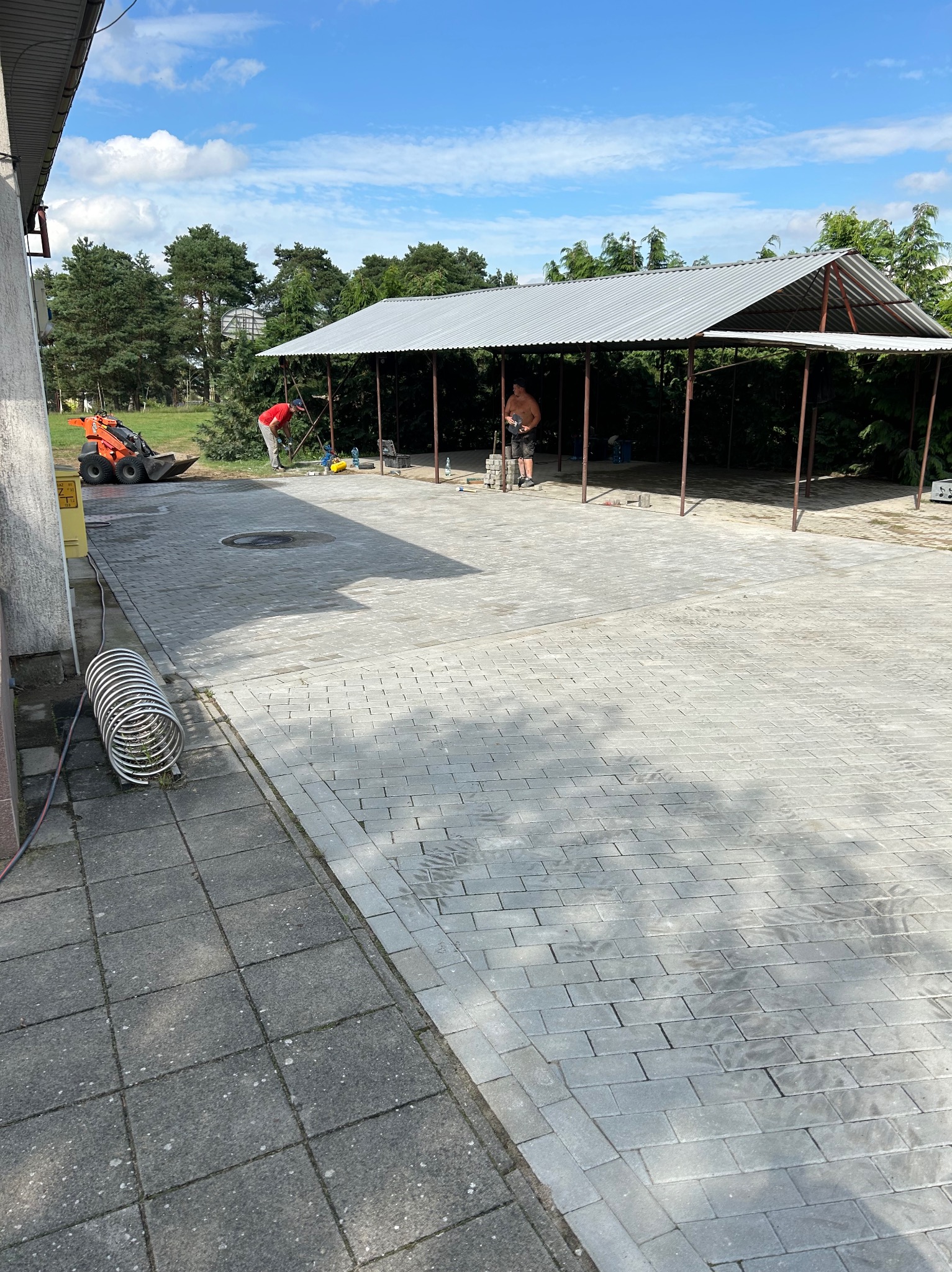 Układanie szarej kostki brukowej na posesji, widoczny mini-ładowarka i pracownicy w trakcie robót.