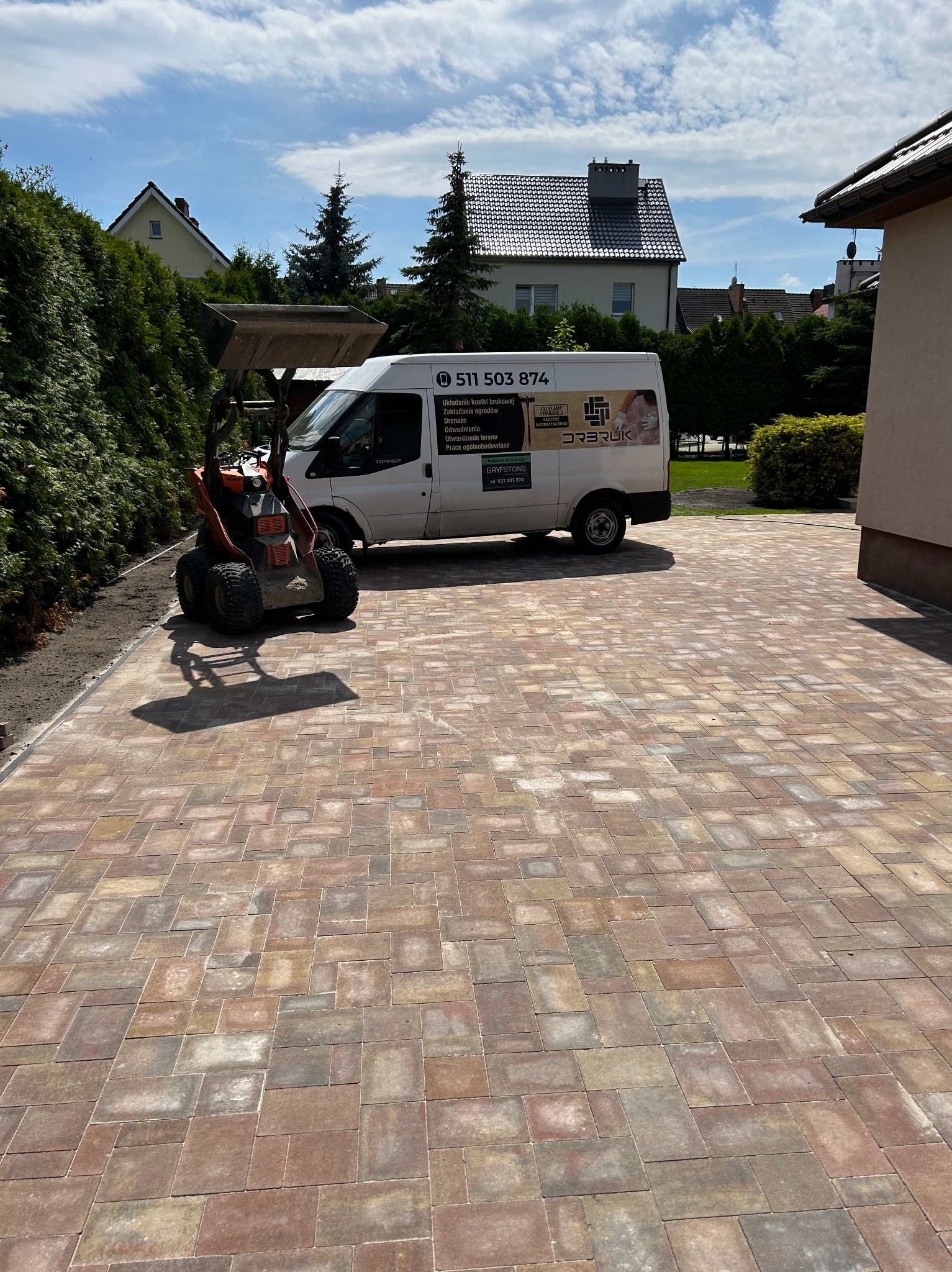 Ułożona kostka brukowa na podjeździe, z zaparkowanym białym busem z reklamą firmy brukarskiej i małą koparką w tle.