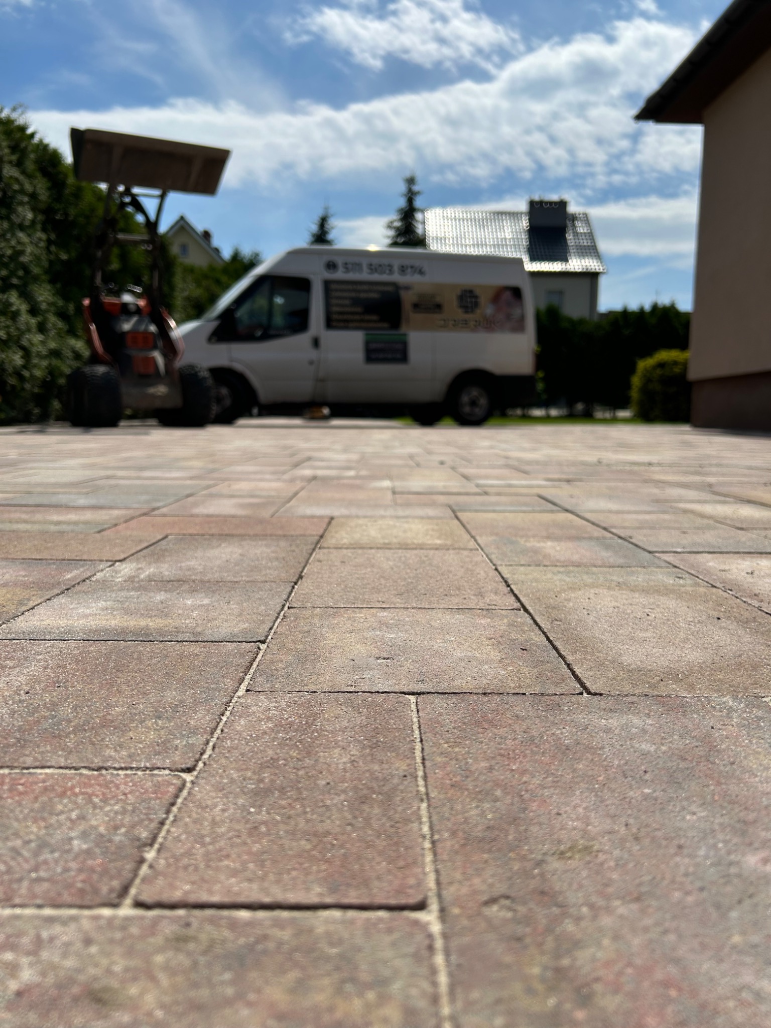 Ułożona kostka brukowa w odcieniach brązu i beżu, widok z niskiej perspektywy, na drugim planie biały bus firmy i sprzęt budowlany, w tle dom jednorodzinny z dachem pokrytym dachówką.