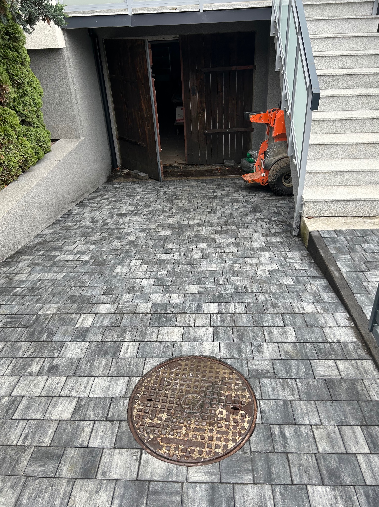 Ułożona szara kostka brukowa prowadząca do otwartych, ciemnych, drewnianych drzwi piwnicy z pomarańczową mini-ładowarką widoczną w środku, studzienka kanalizacyjna na pierwszym planie.