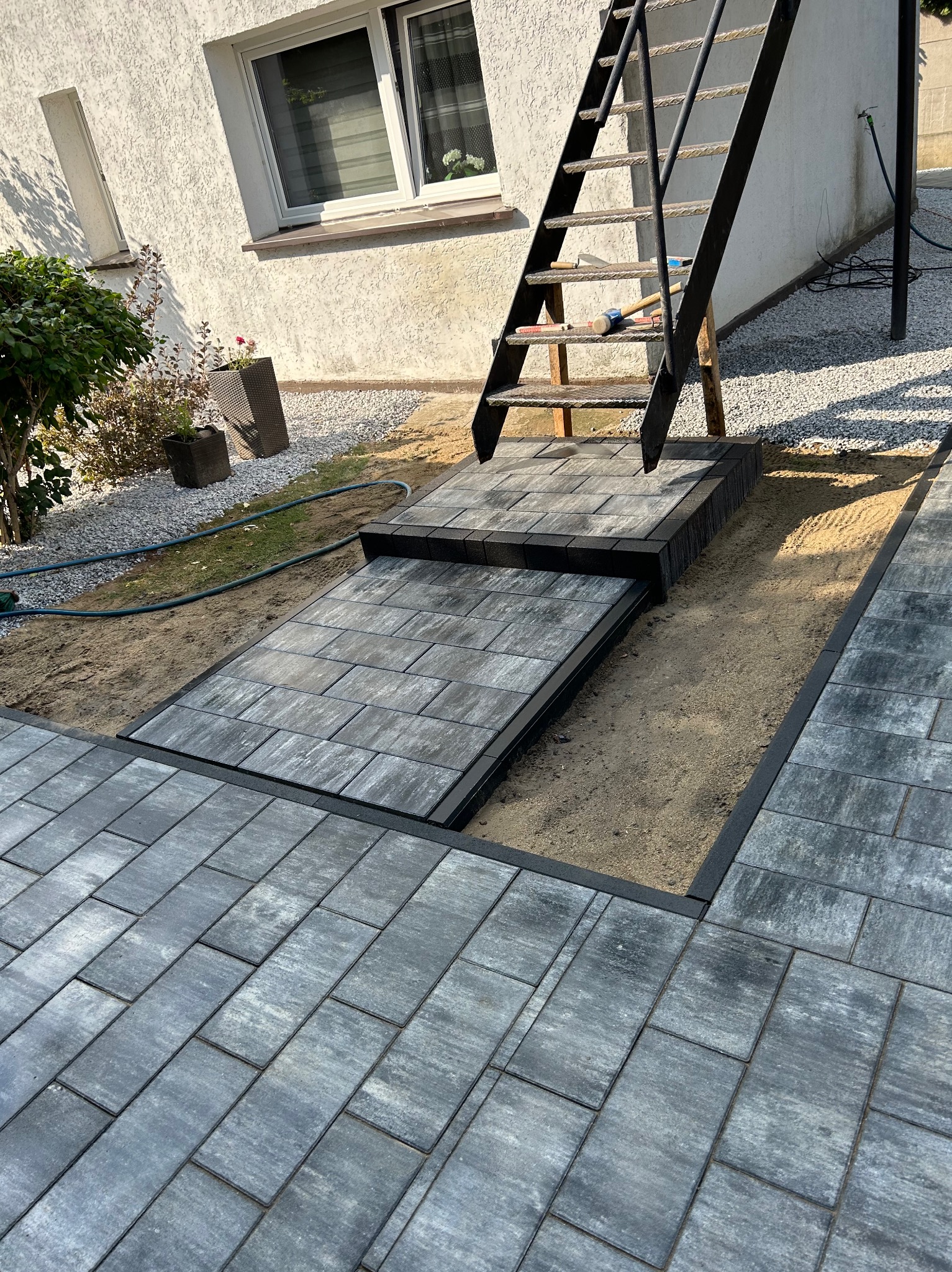 Fragment nowej nawierzchni z szarej kostki brukowej ułożonej przy schodach zewnętrznych, widoczne narzędzia na schodach, obrzeża z czarnego tworzywa.