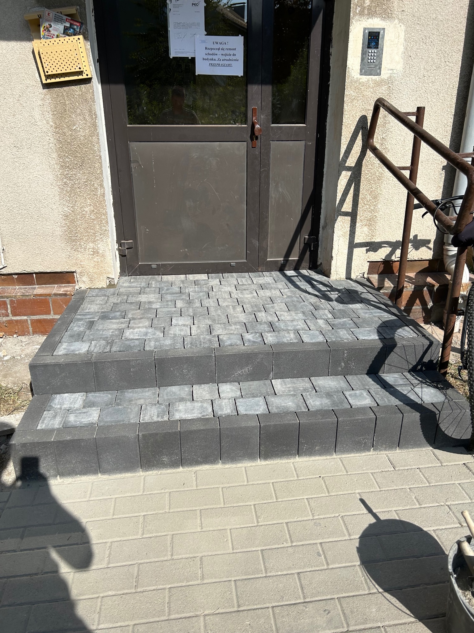 Nowe, dwustopniowe schody wejściowe z szarej kostki brukowej, prowadzące do budynku z brązowymi drzwiami. Na ścianie obok drzwi widoczna skrzynka na listy i domofon. Na drzwiach wisi kartka...