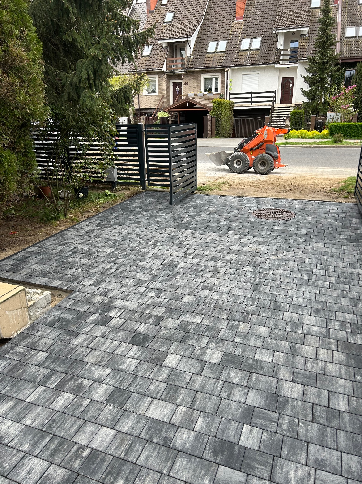 Szara kostka brukowa ułożona na podjeździe, widoczny fragment niezakończonej krawędzi i pomarańczowa miniładowarka w tle, czarne metalowe ogrodzenie po bokach.
