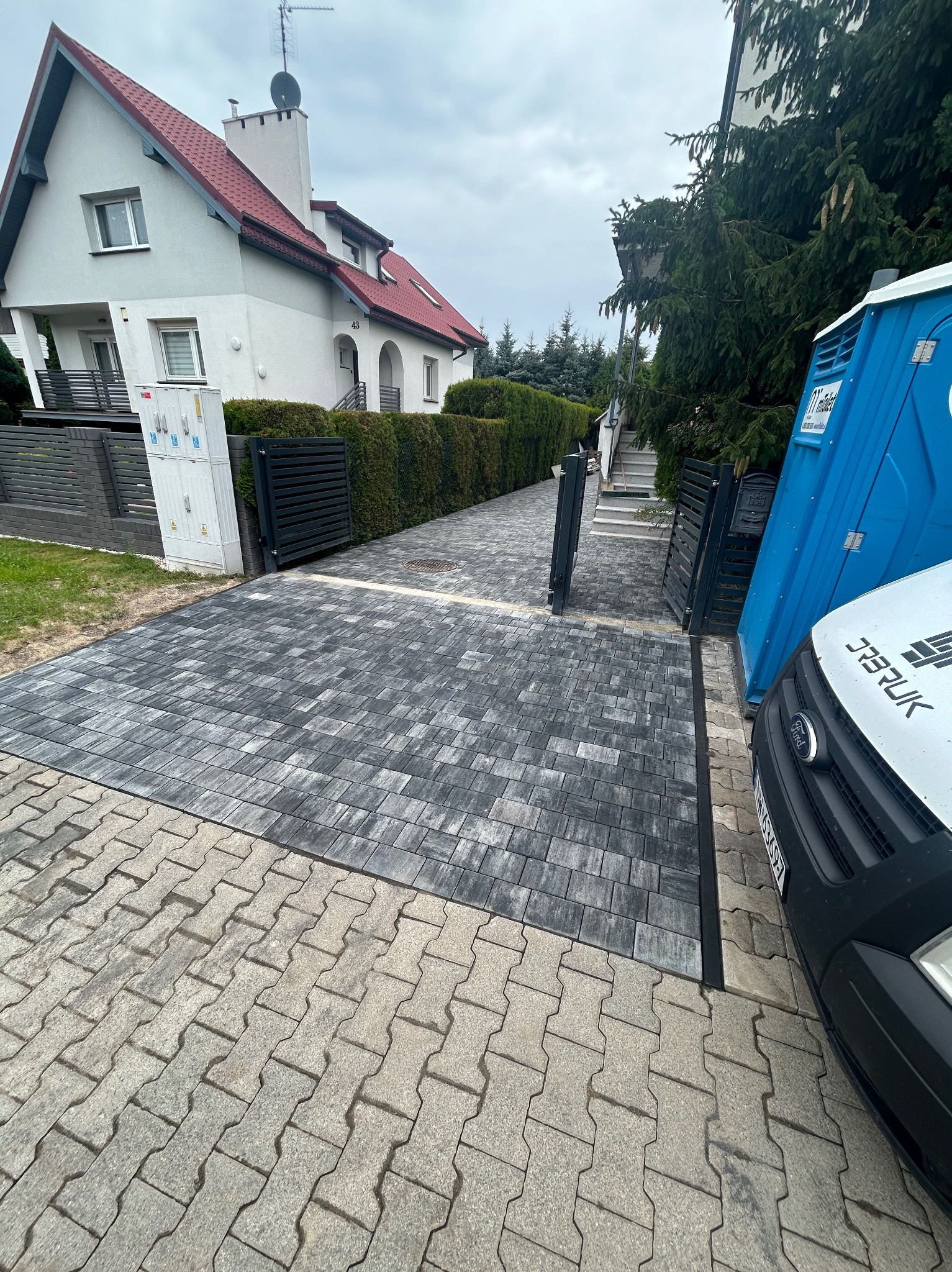 Ułożona, szara kostka brukowa prowadząca do bramy posesji, obok zaparkowany biały samochód dostawczy i toaleta przenośna.