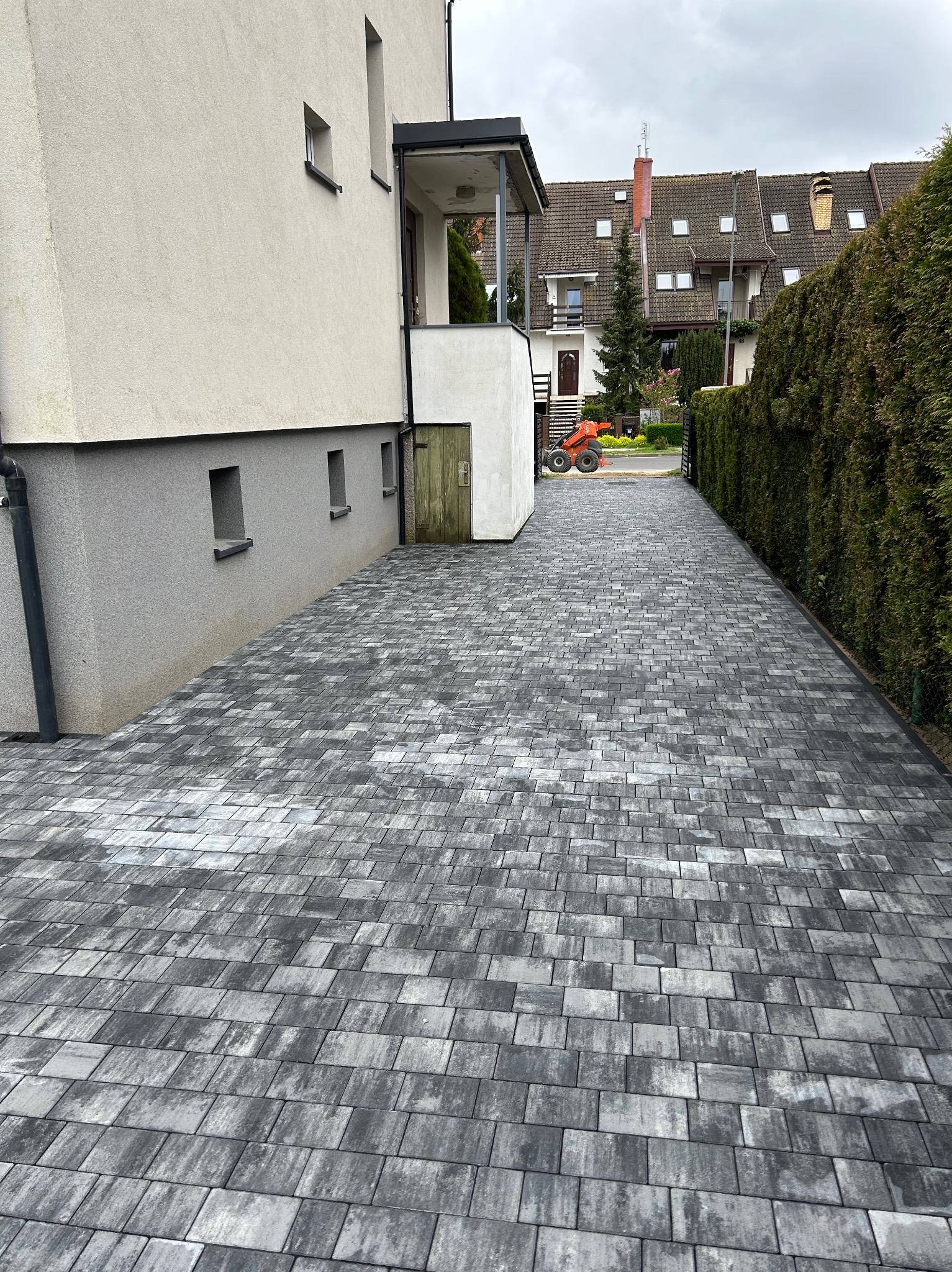 Ułożona szara kostka brukowa na podjeździe, widok z boku domu, w tle pomarańczowa miniładowarka i zabudowa szeregowa.