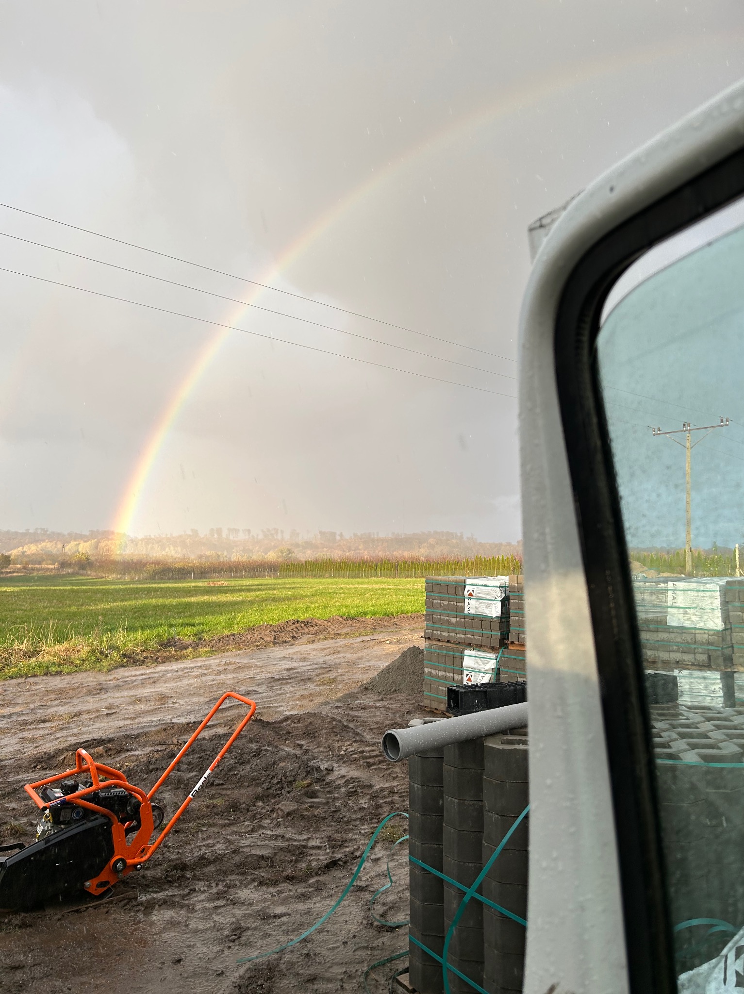 Pomarańczowa zagęszczarka w błocie, stosy szarych kostek brukowych w tle, widok z kabiny samochodu na pole i podwójną tęczę na pochmurnym niebie.