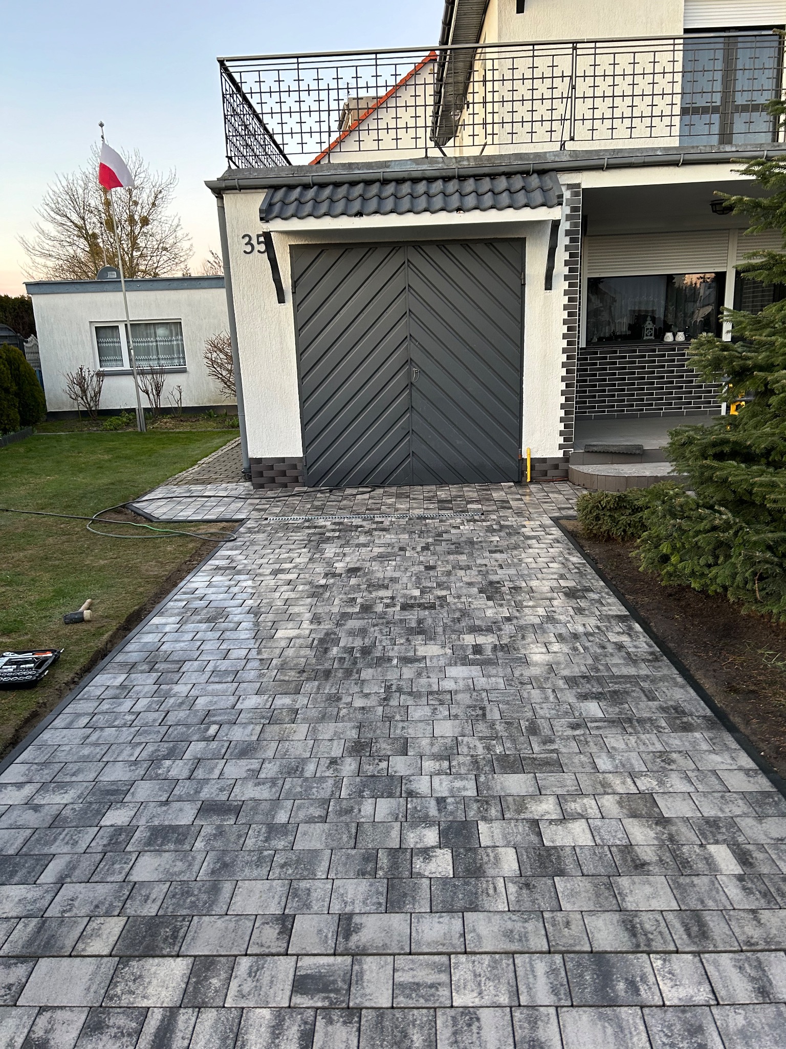 Ułożona szara kostka brukowa prowadząca do garażu domu jednorodzinnego z numerem 35, widoczny polski maszt flagowy i narzędzia brukarskie na trawniku.