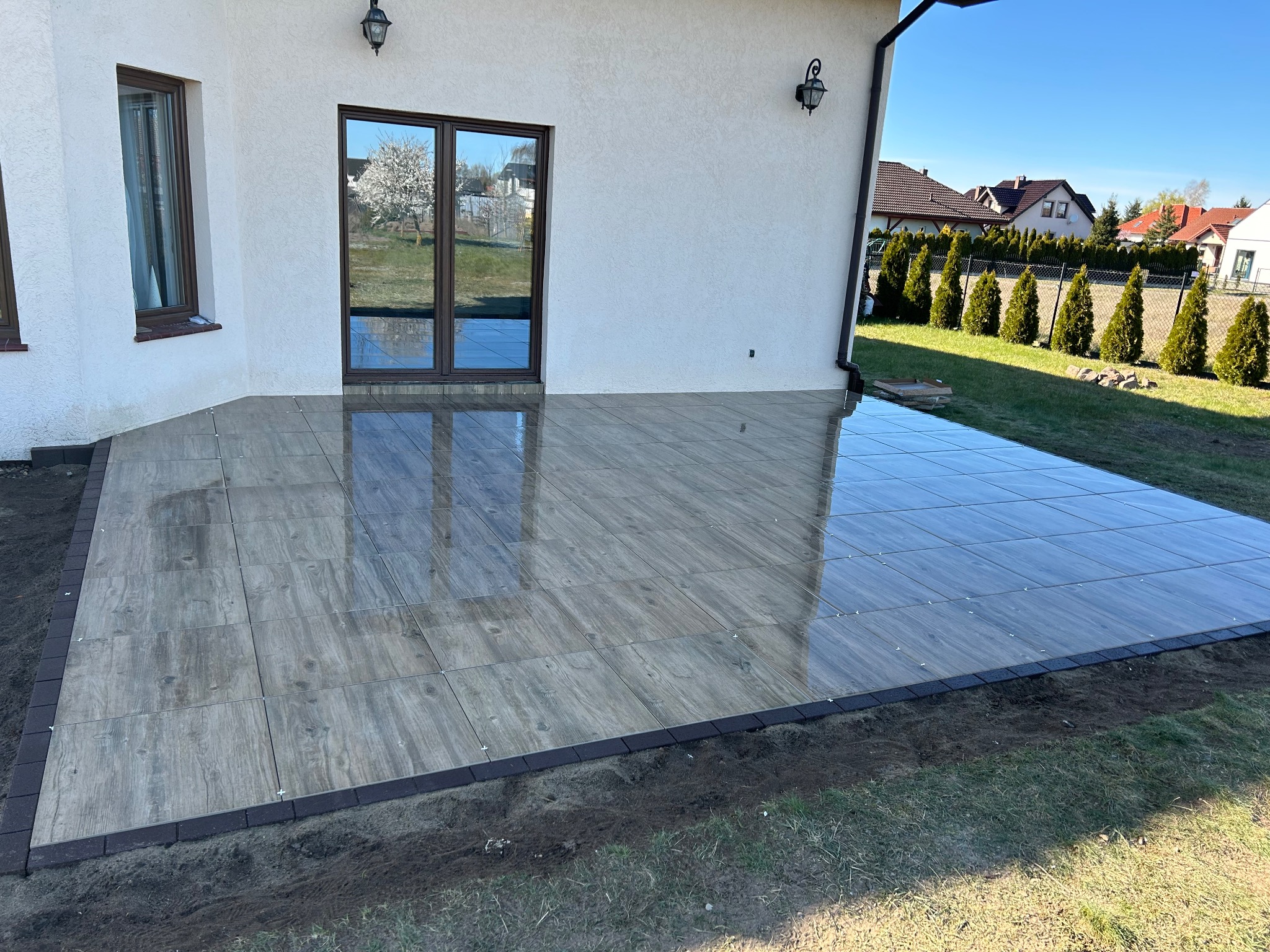 Nowo ułożony taras z płytek imitujących drewno, obramowany ciemną kostką, z widocznym odbiciem kwitnącego drzewa w szklanych drzwiach tarasowych.