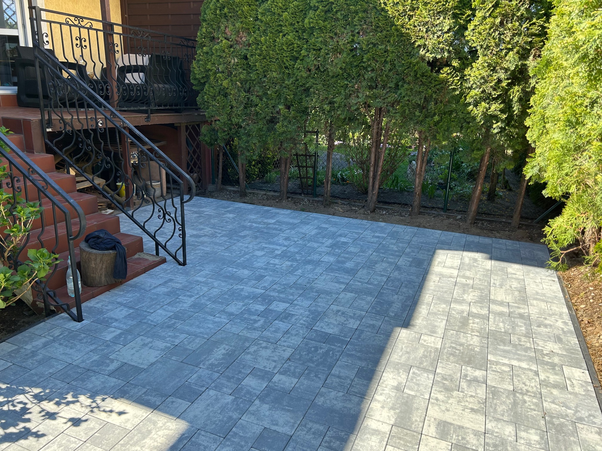 Ułożona szara kostka brukowa na podwórku z zielonymi tujami w tle. Widoczne schody z ozdobną, metalową balustradą. Na schodach leży czarna bluza.