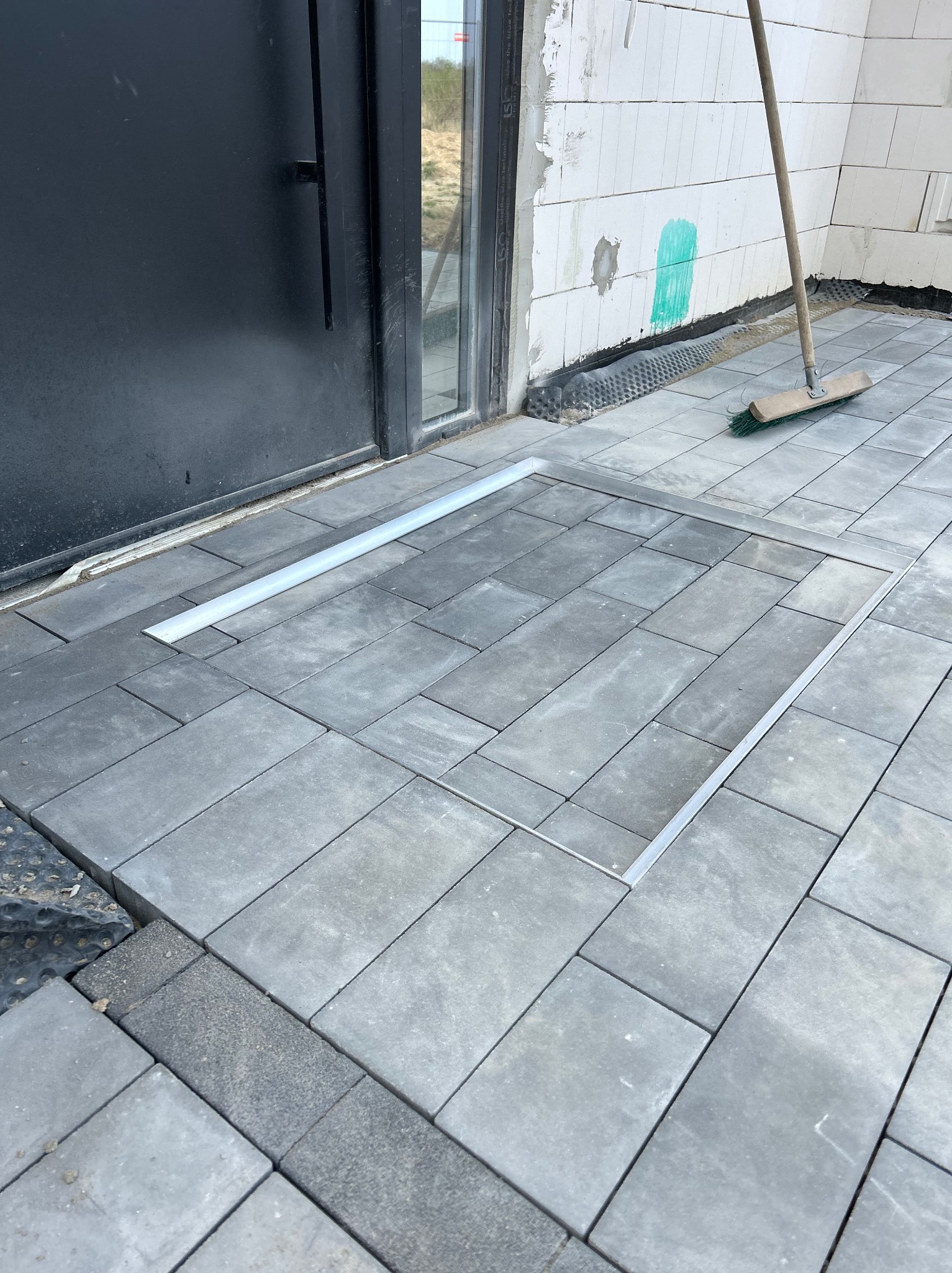 Szara kostka brukowa ułożona przy drzwiach z aluminiową ramką, widoczna szczotka oparta o ścianę z pustaków. Nowoczesne wykończenie.