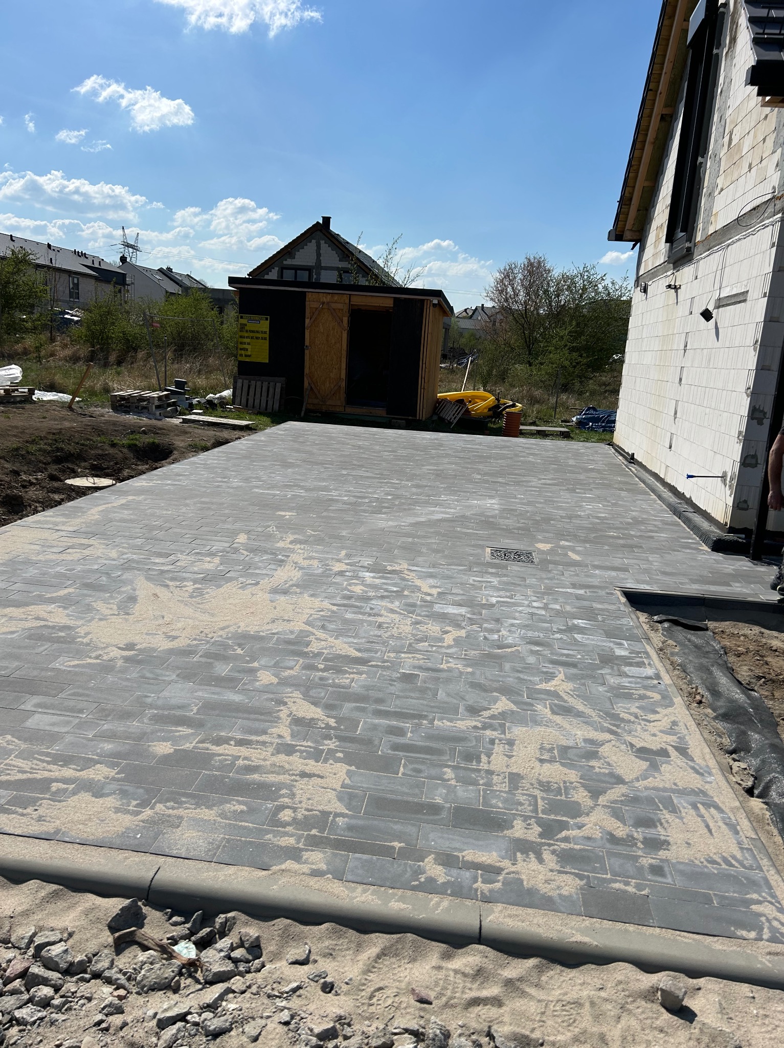 Świeżo ułożona, szara kostka brukowa na posesji, z piaskiem wypełniającym szczeliny. Widoczne obrzeża chodnikowe i fragment budynku w tle, w słoneczny dzień.