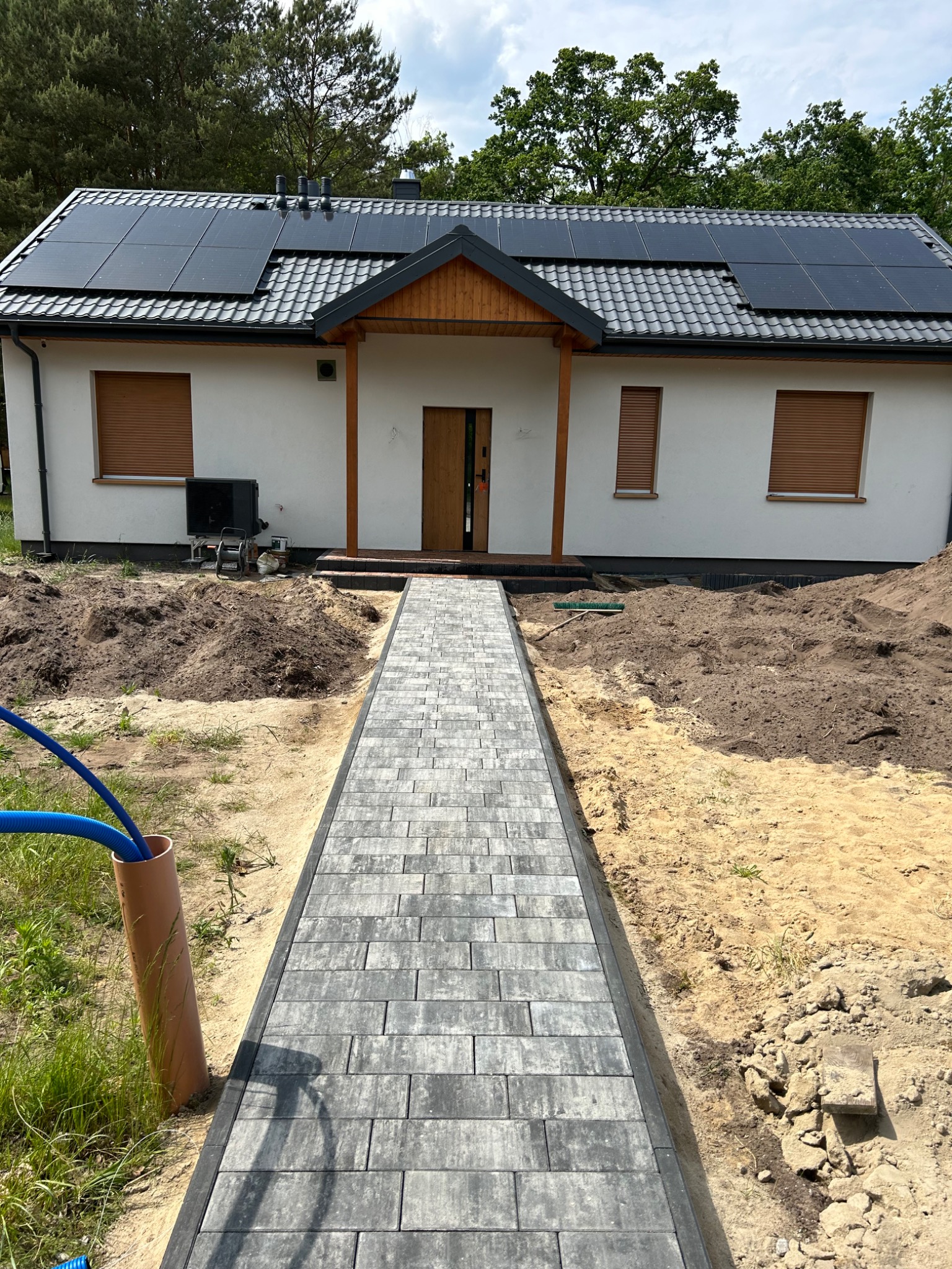 Ułożony chodnik z kostki brukowej prowadzi do nowoczesnego domu z panelami słonecznymi na dachu. Widoczne prace ziemne wokół, rury doprowadzone do budynku.