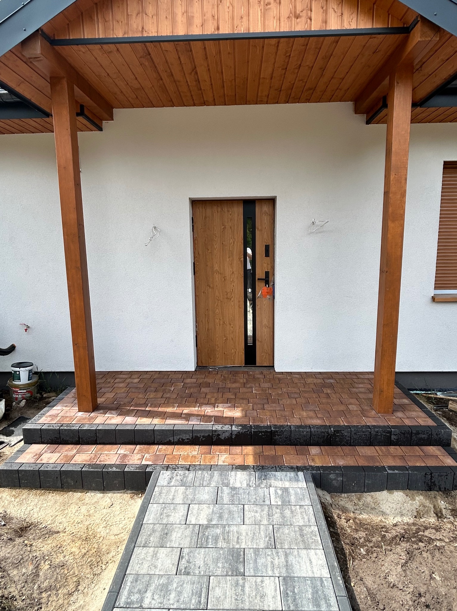 Bruk z kostki w odcieniach brązu prowadzi do wejścia domu z drewnianym zadaszeniem. Schody wykończone ciemną kostką. Częściowo ułożona ścieżka z szarej kostki.