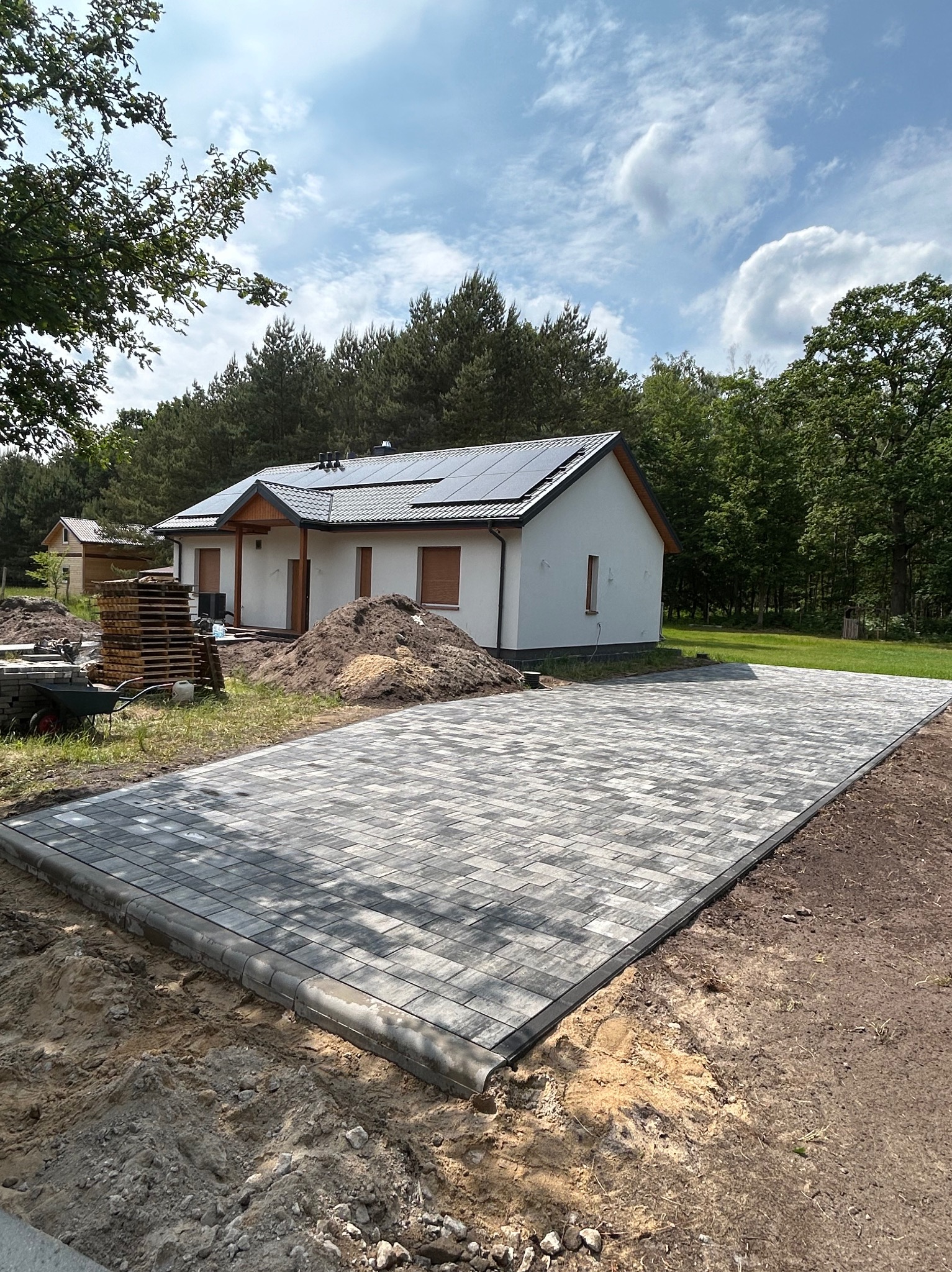 Ułożona, szara kostka brukowa przed domem z panelami słonecznymi. Widoczny fragment ogrodu i drzewa. Ujęcie z poziomu gruntu, z bliska na kostkę.