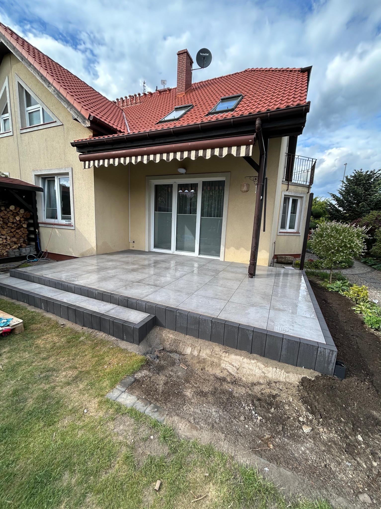 Wykończony taras z szarych płytek ceramicznych przy beżowym domu z czerwonym dachem. Widoczne schody z ciemnych kostek i fragment ogrodu.