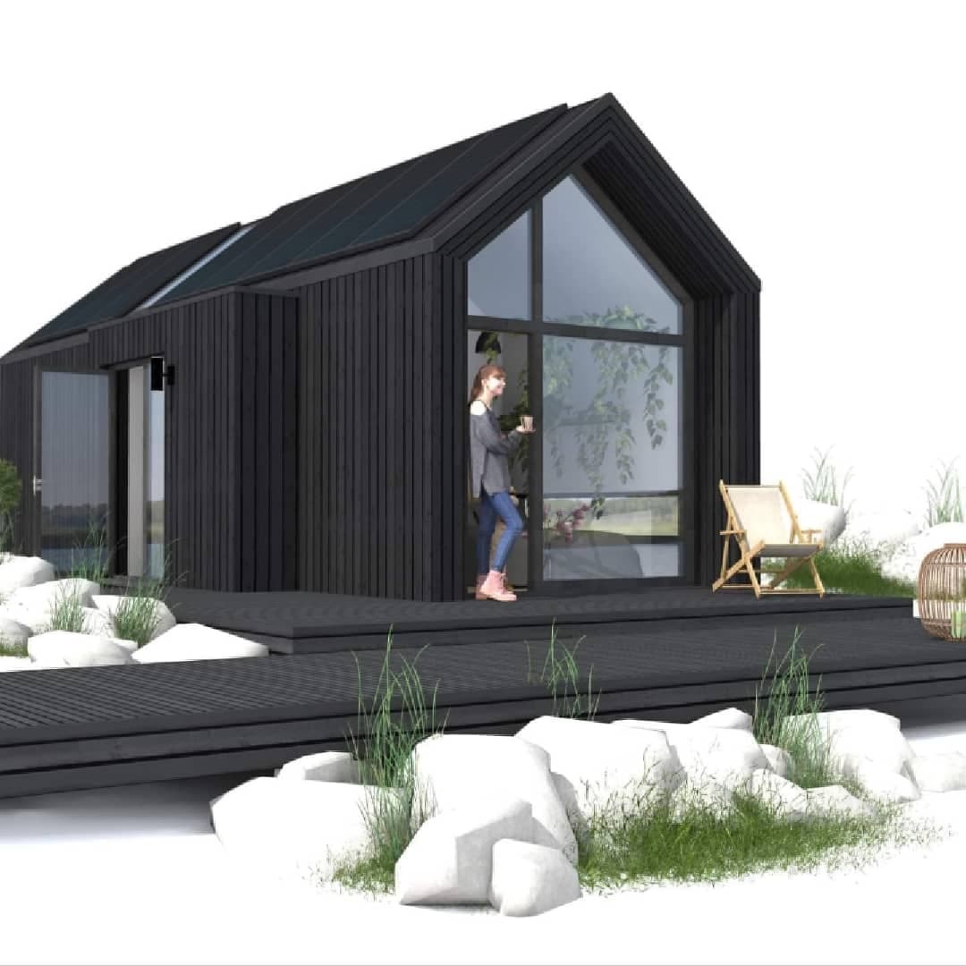 Czarna, nowoczesna bryła domu z panelami słonecznymi na dachu, duże okno szczytowe, kobieta z kubkiem w drzwiach, drewniany taras i białe kamienie w otoczeniu.