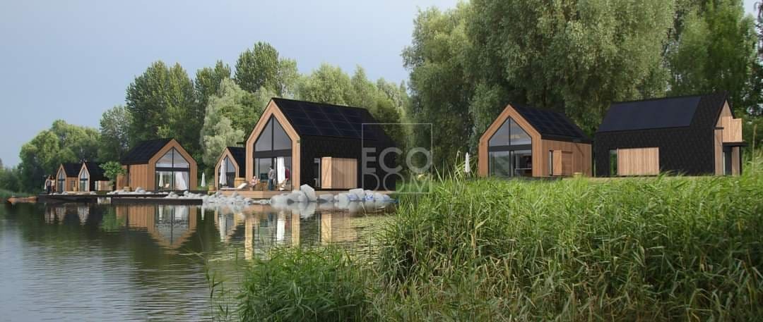 Rząd nowoczesnych, drewnianych domków letniskowych z panelami słonecznymi na dachu, unoszących się na wodzie jeziora, otoczonych zielenią.
