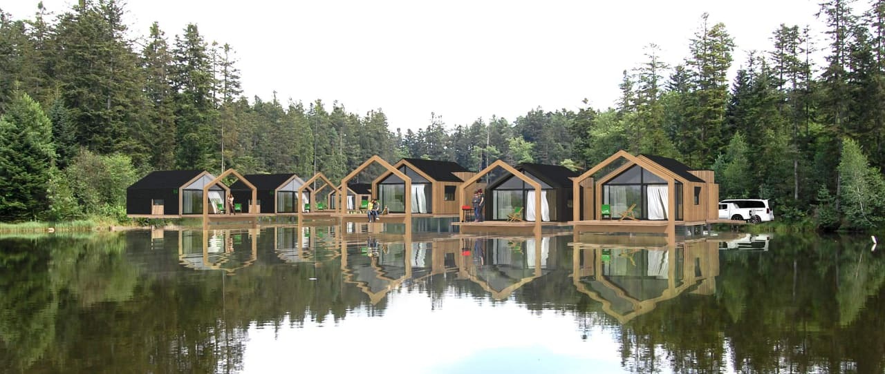 Rząd pływających domków z drewnianą elewacją i czarnymi dachami, odbijających się w spokojnej wodzie jeziora otoczonego lasem.