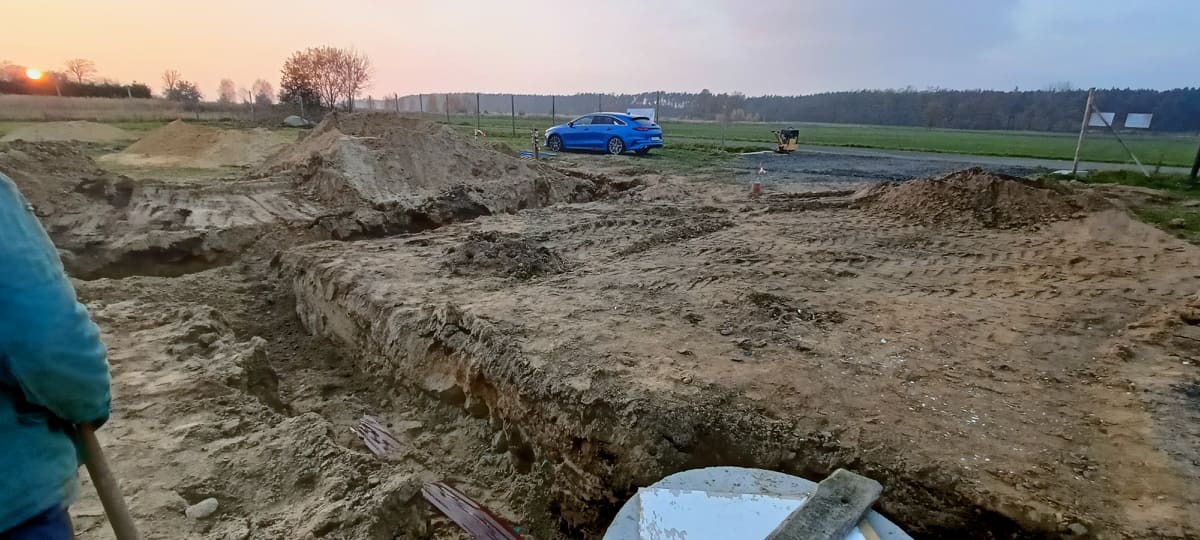 Wykop pod fundamenty domu szkieletowego, widoczna ziemia, wykopane rowy, w tle niebieski samochód osobowy i zachodzące słońce.
