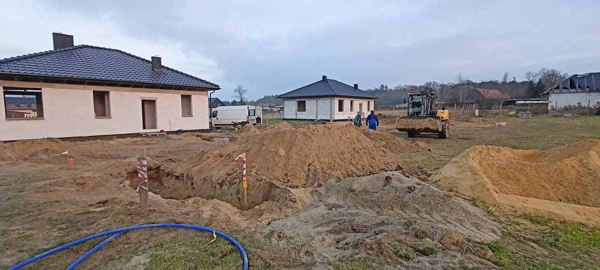 Dwa domy w stanie surowym otwarte w technologii szkieletowej, wykopany fundament, koparka i pracownicy na placu budowy, pochmurne niebo.