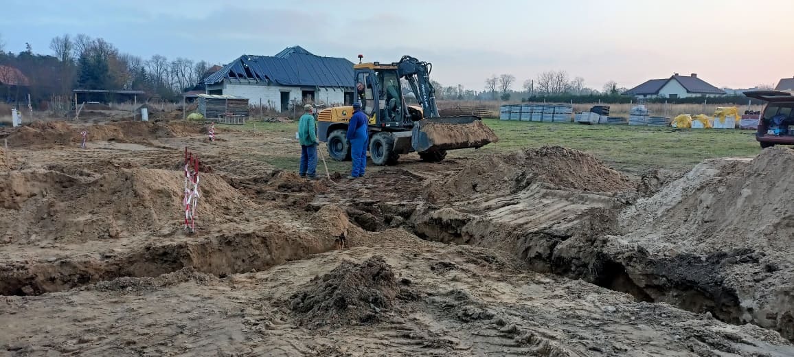 Wykopy pod fundamenty, dwóch robotników i koparka na placu budowy, widoczne materiały budowlane w tle.