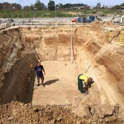 Wykop pod zbiornik betonowy z pracownikami w środku, widoczne rury na obrzeżach i sprzęt budowlany w tle. Prace ziemne w toku, słoneczny dzień.