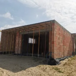 Dom murowany w trakcie budowy, ściany z czerwonej cegły, widoczne podpory stropu, teren wokół przygotowany pod dalsze prace.