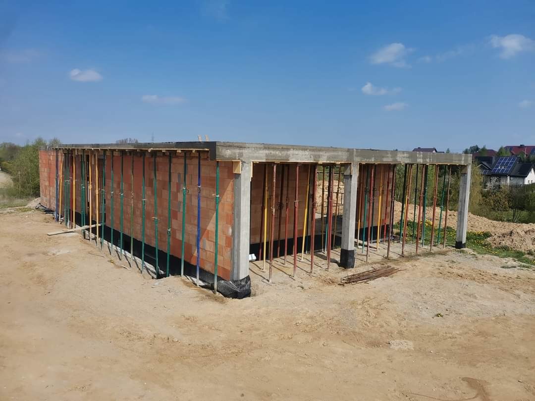 Wznoszenie ścian z czerwonej cegły na fundamencie z izolacją przeciwwilgociową, wsparte stalowymi stemplami w kolorach zielonym, żółtym i niebieskim, na tle błękitnego nieba i zabudowań mieszkalnych.