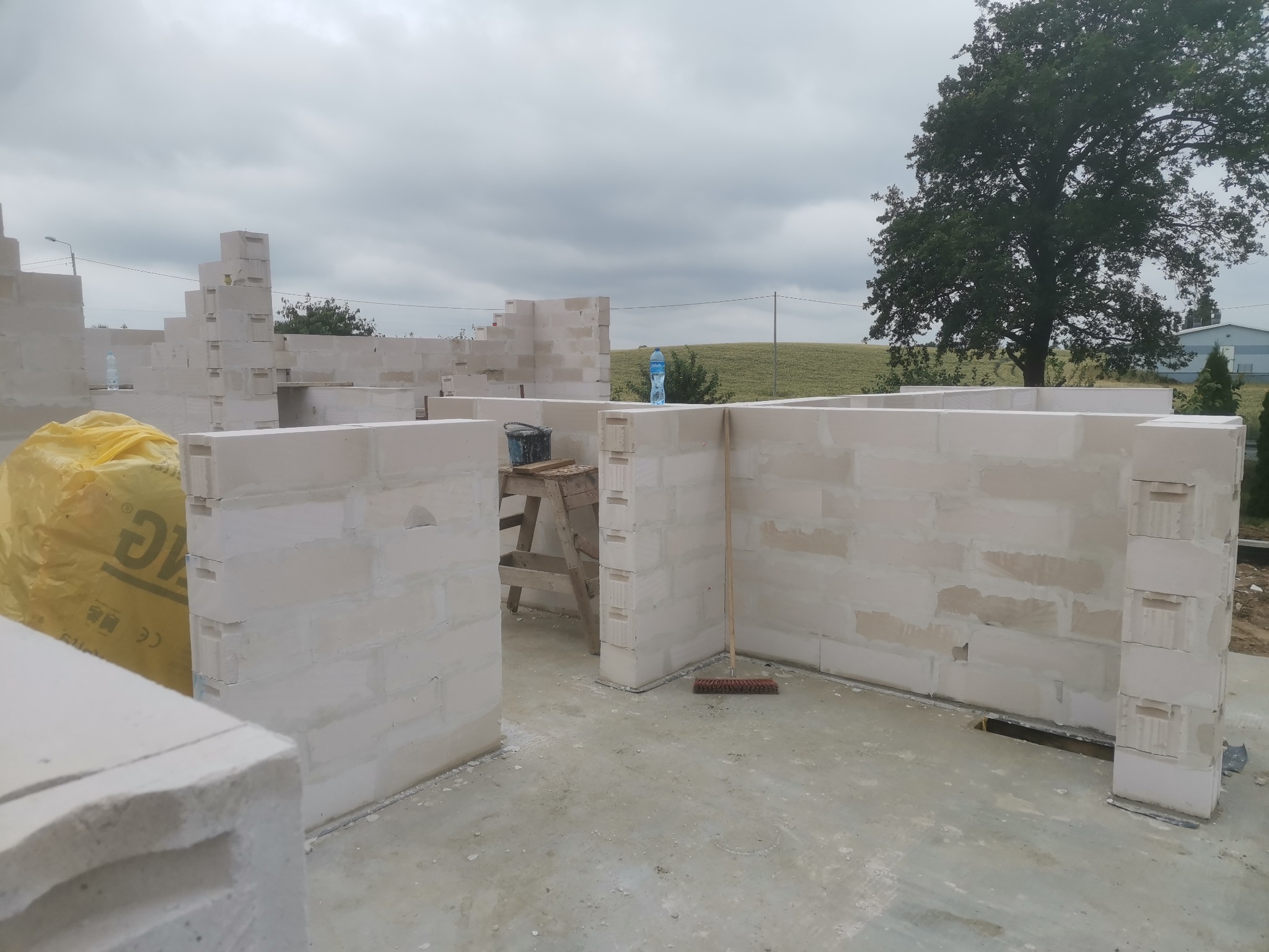 Wznoszone ściany domu z bloczków betonu komórkowego, widoczne rusztowanie, wiadro i butelka wody, na zewnątrz krajobraz polny.