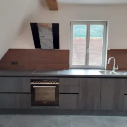 Nowoczesna, szara zabudowa kuchenna pod skosem dachu, z oknem wychodzącym na zieleń, z płytą grzewczą i piekarnikiem, oraz drewnianym panelem nad blatem.