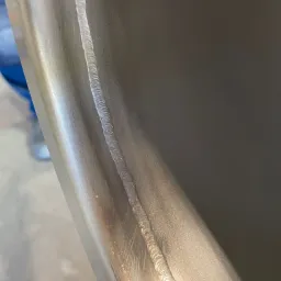 Metalowa powierzchnia z wyraźnym, równym spoiwem spawalniczym biegnącym wzdłuż krawędzi. Widoczne ślady obróbki i delikatne refleksy światła na metalu.